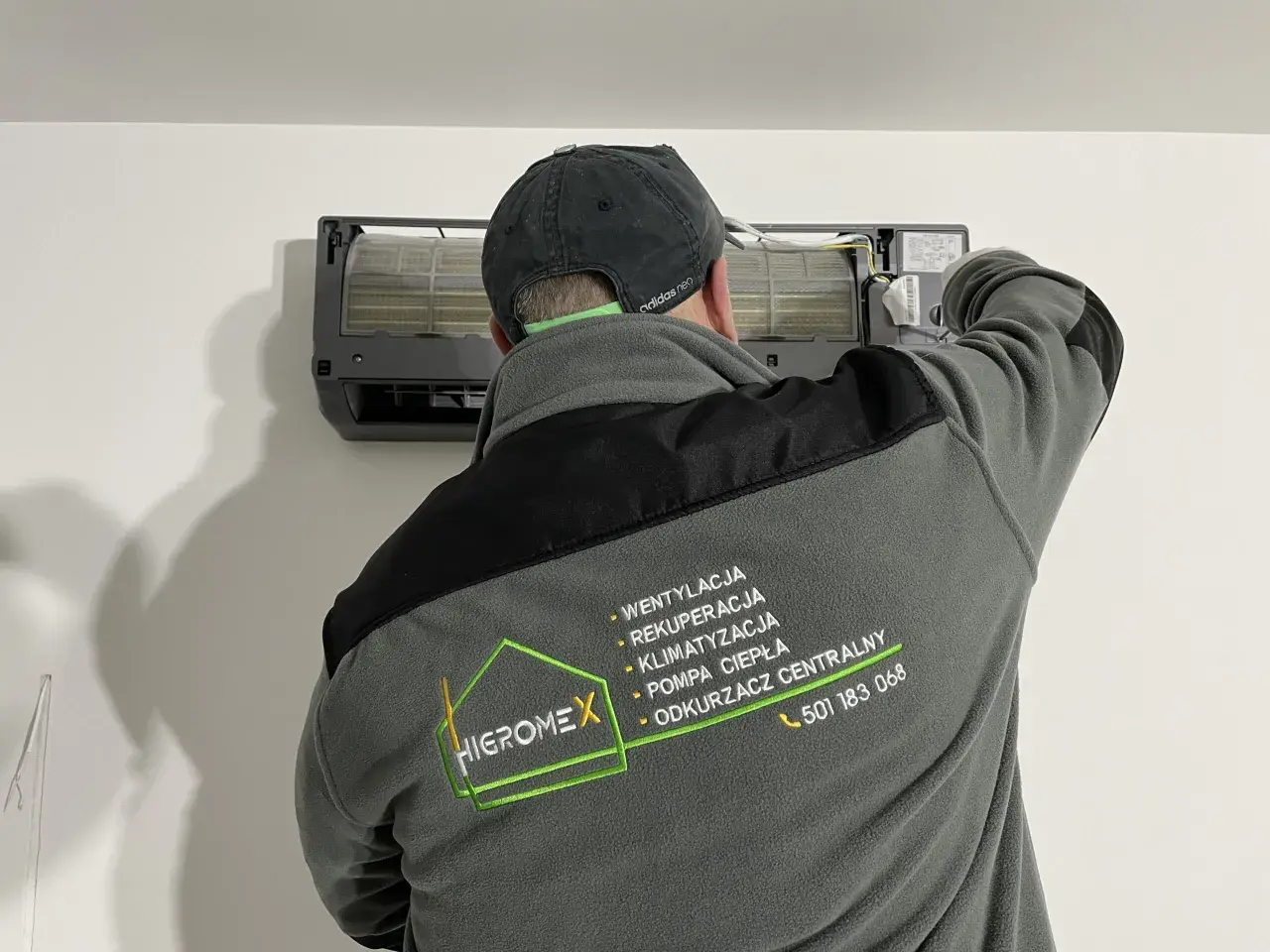 Technik w czapce z daszkiem montuje klimatyzator. Na kurtce logo firmy Higromex.