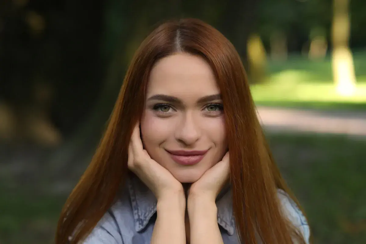 Uśmiechnięta szatynka z rudymi włosami opiera podbródek na dłoniach, patrząc w obiektyw.