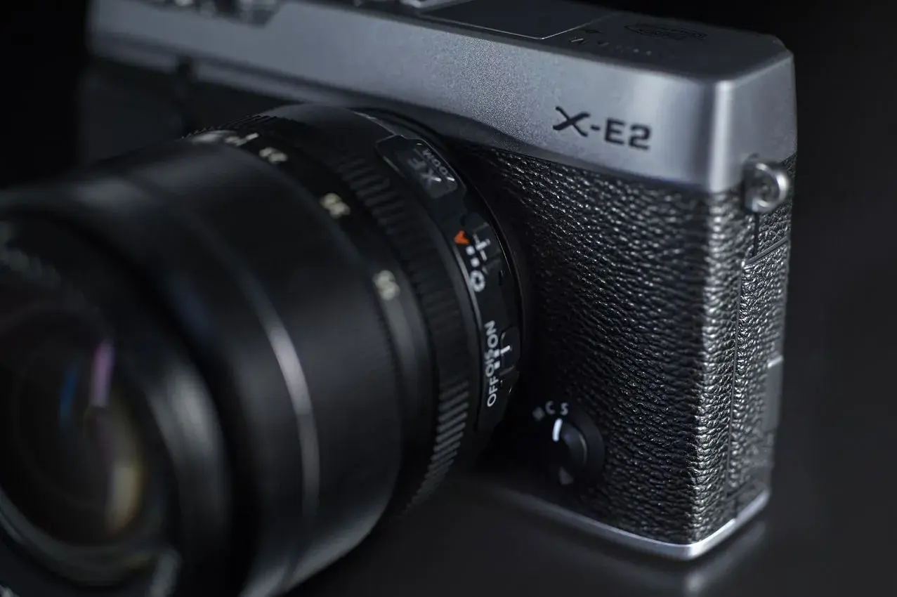 Srebrny aparat Fujifilm X-E2 z obiektywem. Widoczny napis "X-E2" i pokrętło wyboru trybu.