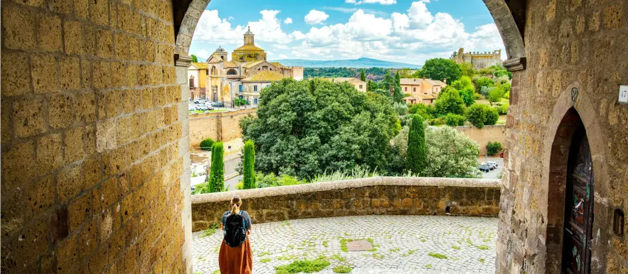 A traveler pauses, backpack ready, gazing at the historic Italian landscape. This view, perhaps on a journey from Viterbo to Rome, showcases ancient architecture and lush greenery under a bright sky.