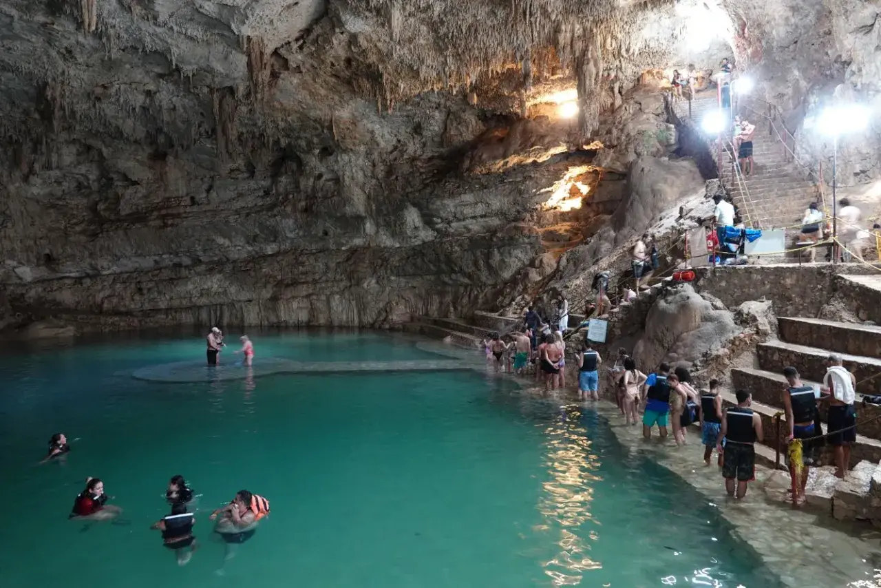 Menschen schwimmen und stehen im türkisfarbenen Wasser einer Höhle in Valladolid, Mexiko. Treppen führen zu einer Plattform, auf der sich weitere Besucher aufhalten.