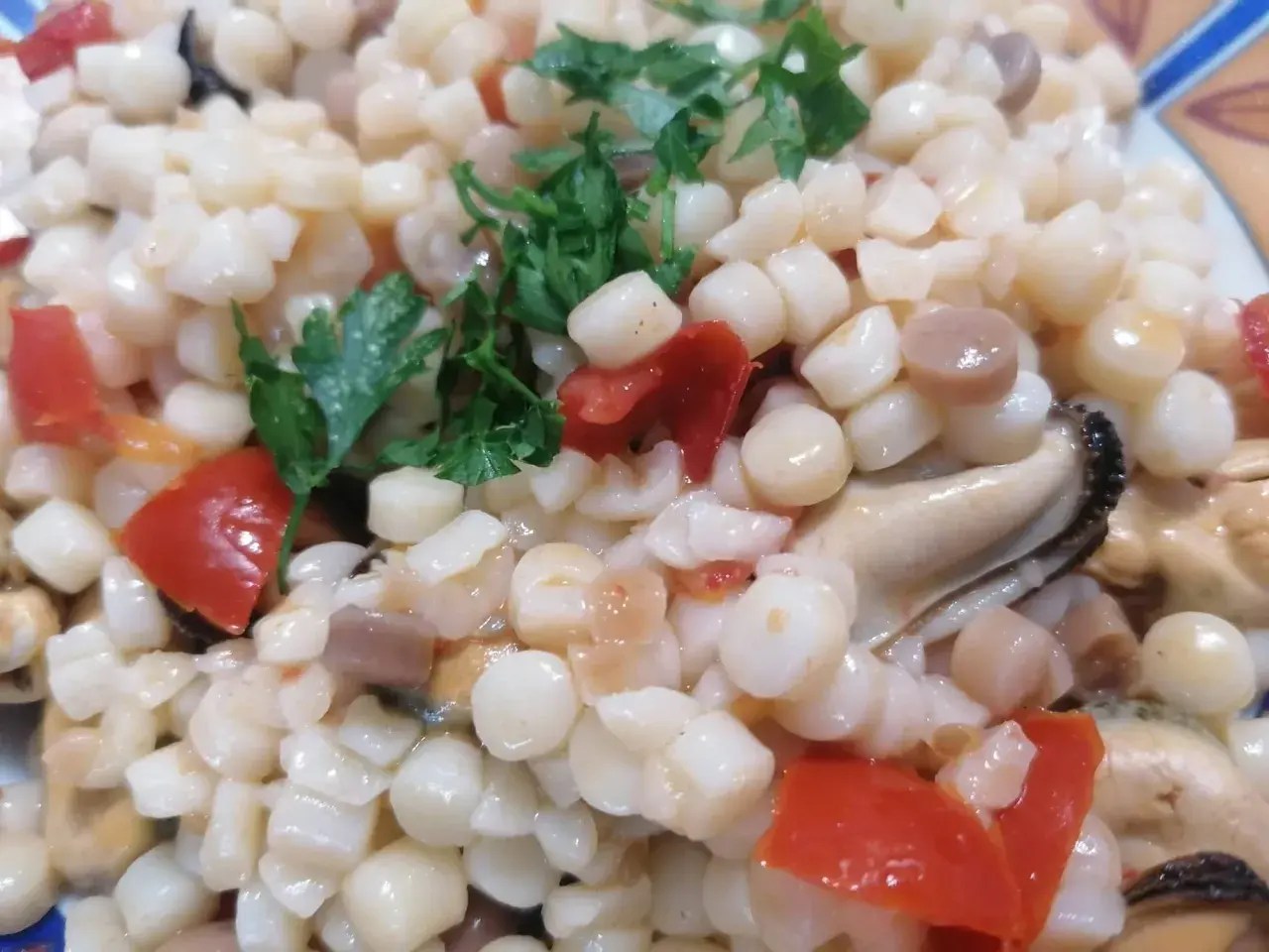 En närbild på en tallrik med fregola pasta, musslor och tärnad tomat, toppad med färsk persilja.