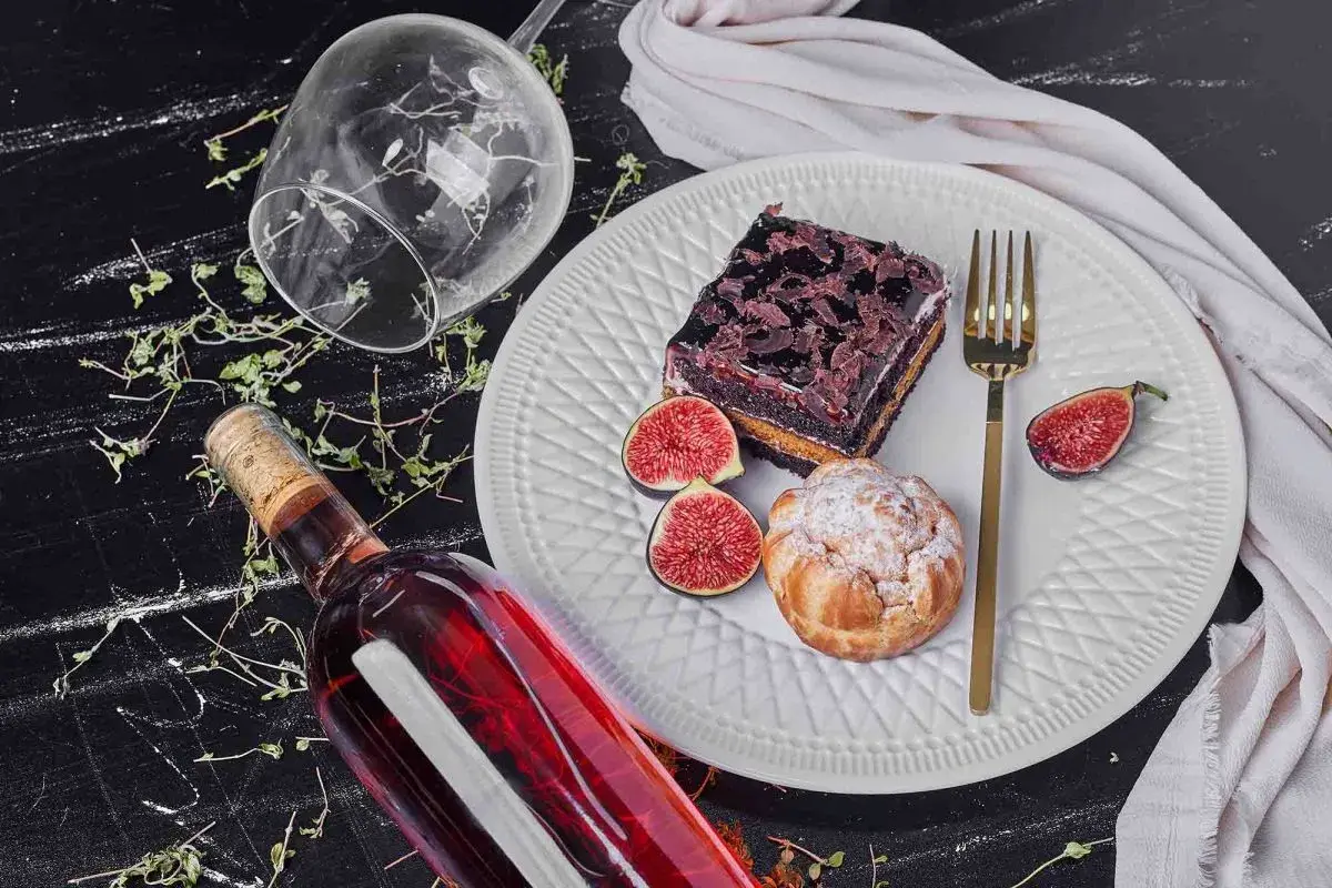 Un bicchiere rovesciato, una bottiglia di vino rosso e un piatto con torta al cioccolato, bignè, fichi e posate dorate, un'invitante composizione per gustare vini da dessert.
