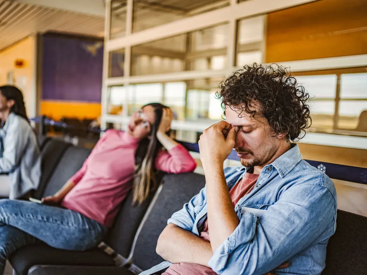 Erschöpfter Mann im Wartebereich, der sich die Augen reibt. Die Frau neben ihm wirkt ebenfalls müde. Gute **Jetlag Tipps** sind hier gefragt.