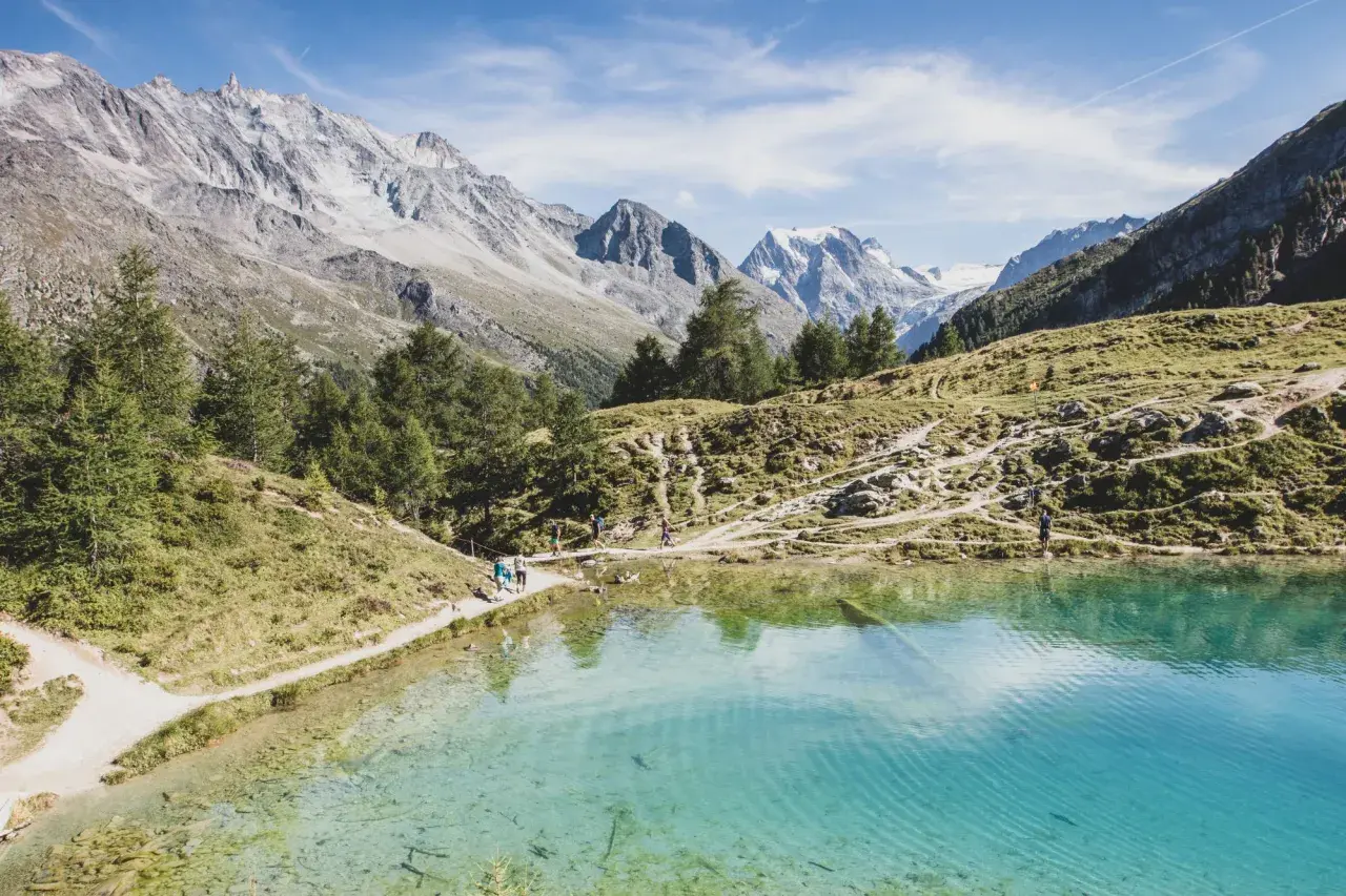 Sentier de randonnée longeant un lac turquoise dans le Val d'Hérens, avec des montagnes majestueuses en arrière-plan.