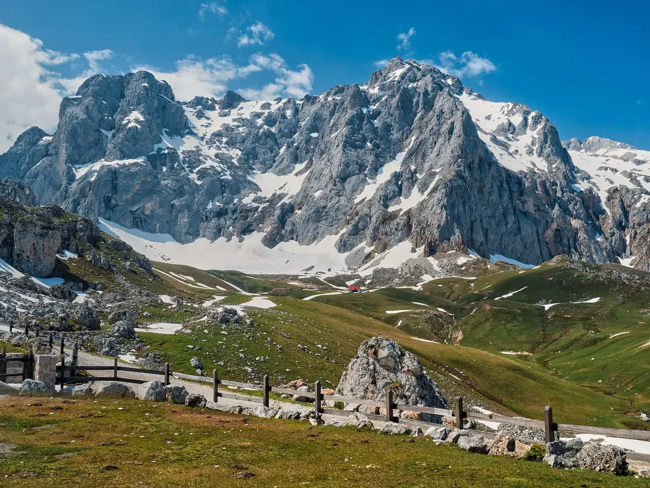 Majestuosas montañas nevadas en los Picos de Europa, cerca de Llanes. Un camino de piedra y madera serpentea por prados verdes salpicados de nieve.