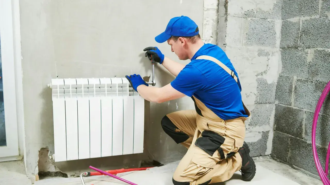 Instalator w niebieskiej czapce i ogrodniczkach pracuje przy grzejniku. Pokazuje, jak zdjąć grzejnik, używając klucza.