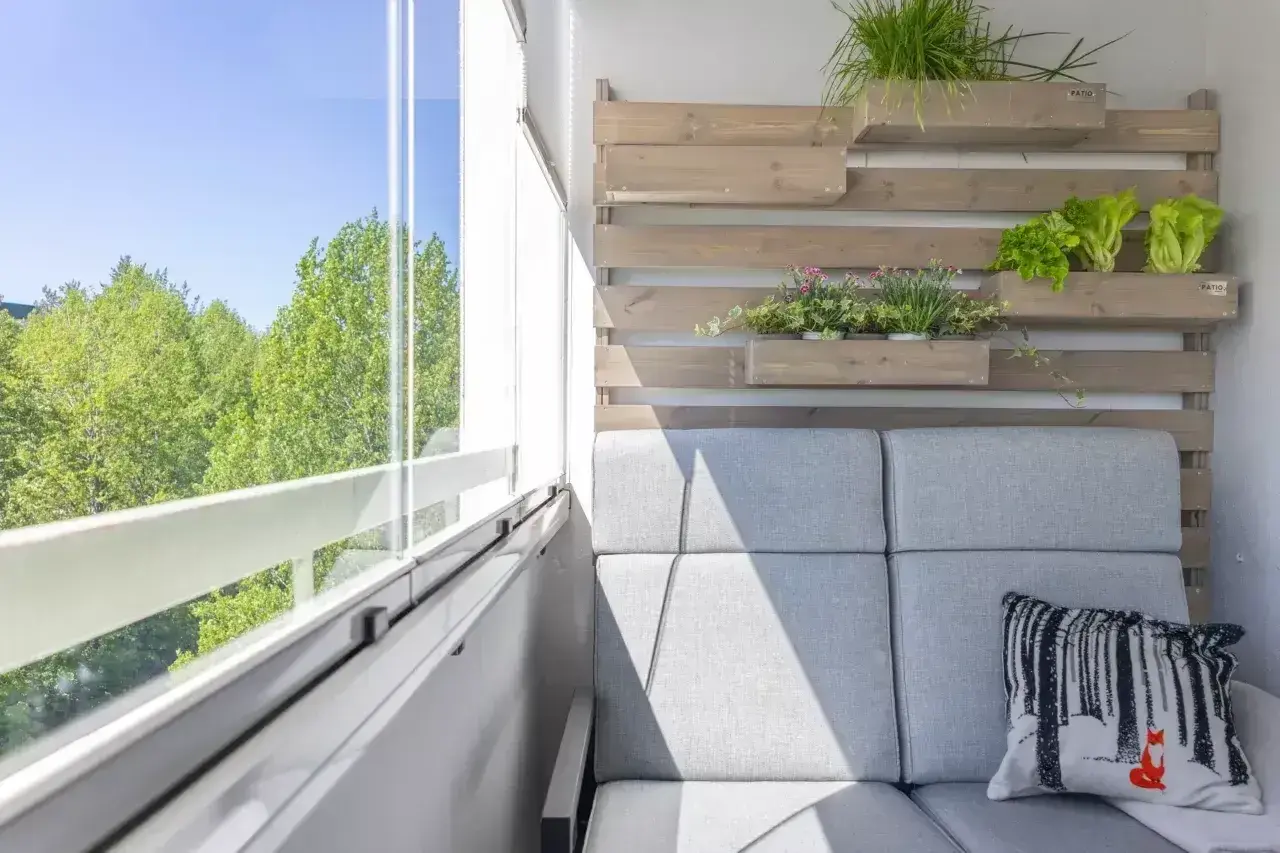 Przytulna loggia z szarą sofą, poduszką z liskiem i zielenią na drewnianej ścianie. Widok na drzewa przez przeszklony balkon.