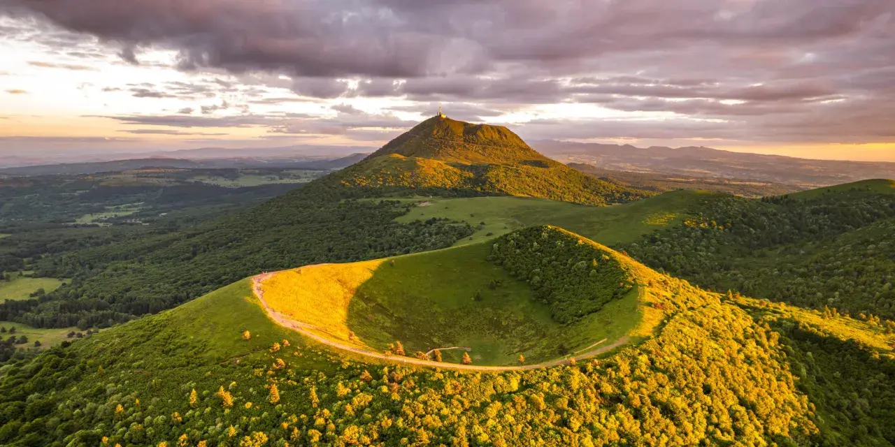 Vue panoramique du Puy de Dôme au coucher du soleil, avec un chemin serpentant sur une colline verdoyante. Idéal pour un séjour en camping-car.