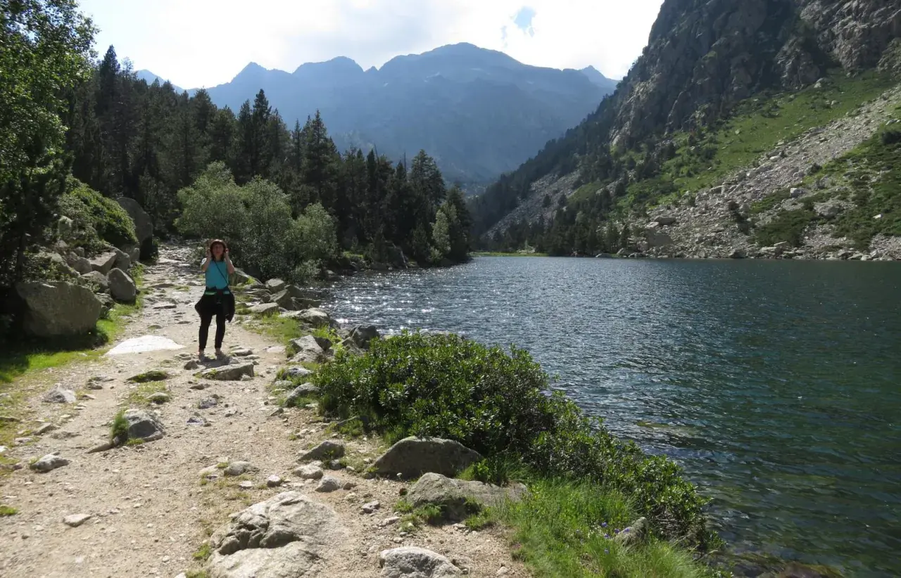 Una mujer camina por un sendero rocoso junto a un estany llong, rodeado de montañas y pinos.