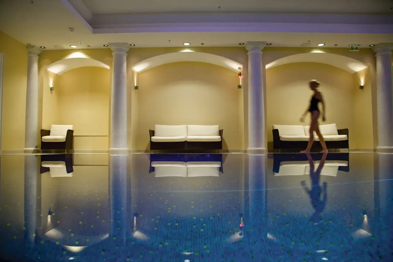 Una mujer camina junto a una piscina interior, rodeada de sofás y columnas, creando una atmósfera de relajación para una experiencia spa sensorial.