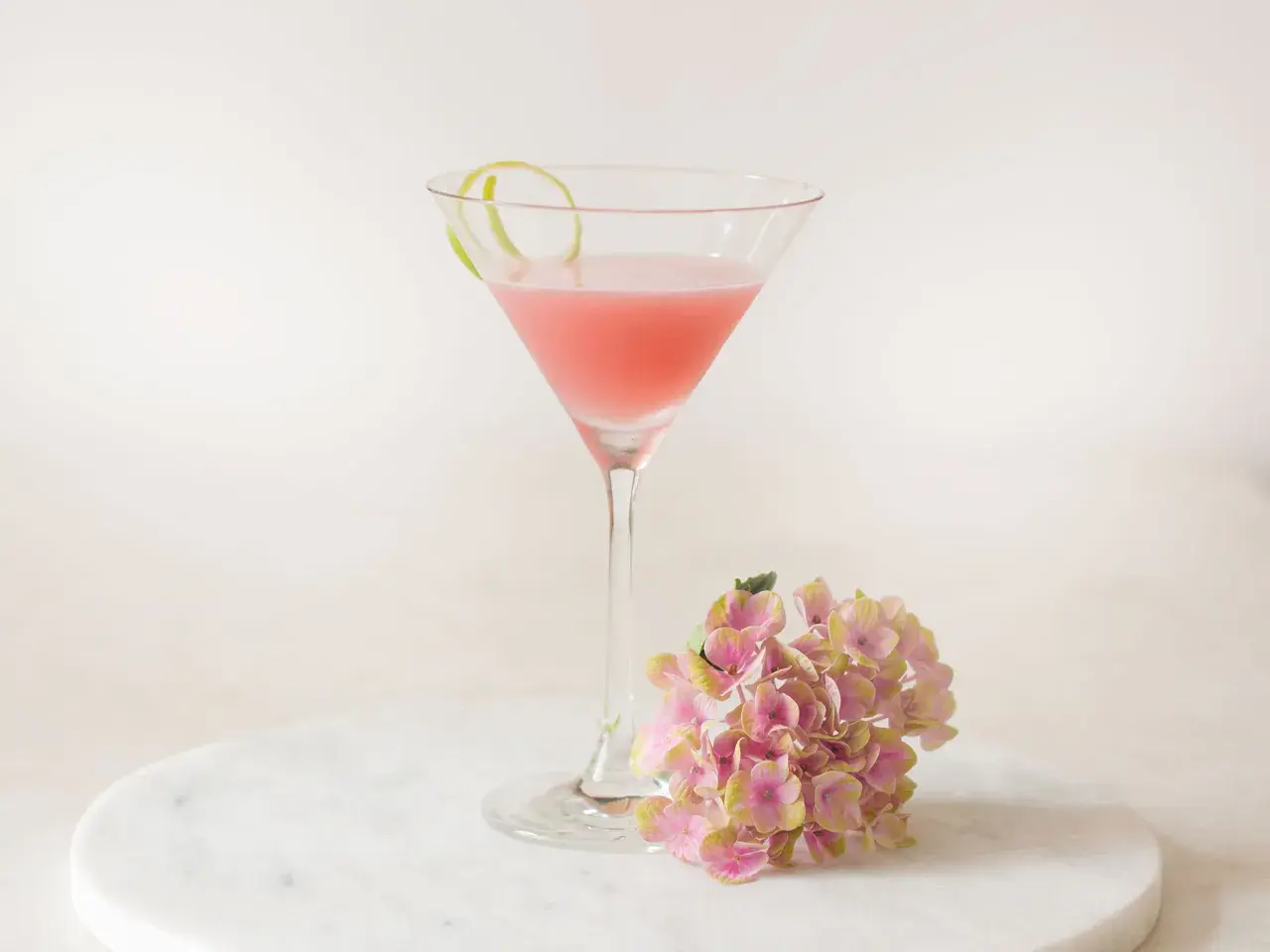 Ein rosafarbener Cosmopolitan Cocktail mit Limettenschale und einer Hortensienblüte auf Marmor.