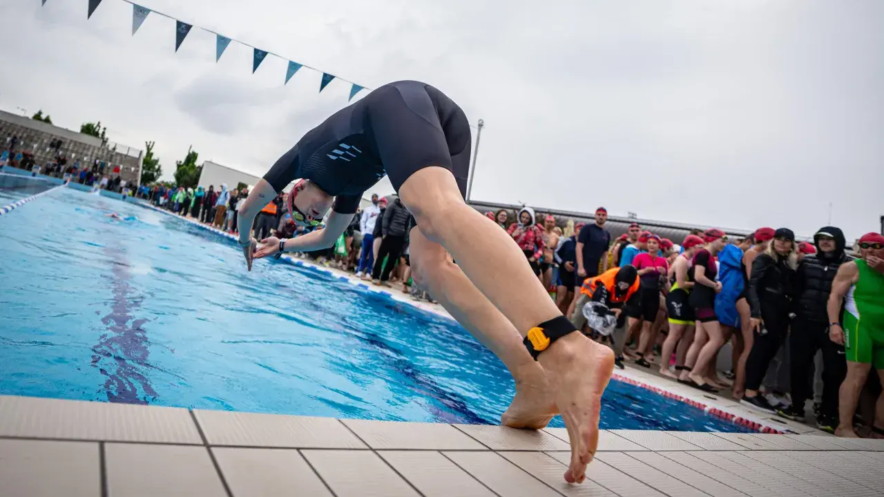 A swimmer in a black wetsuit dives into a pool, ready for advanced swim workouts. Spectators watch from the side.