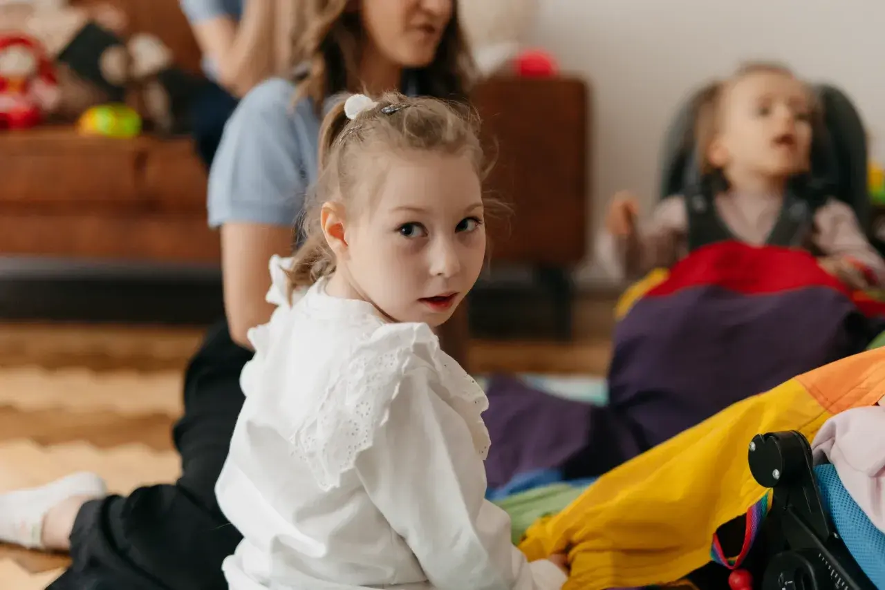 Dziewczynka w białej bluzce patrzy w obiektyw, w tle kobieta i dziecko w wózku.