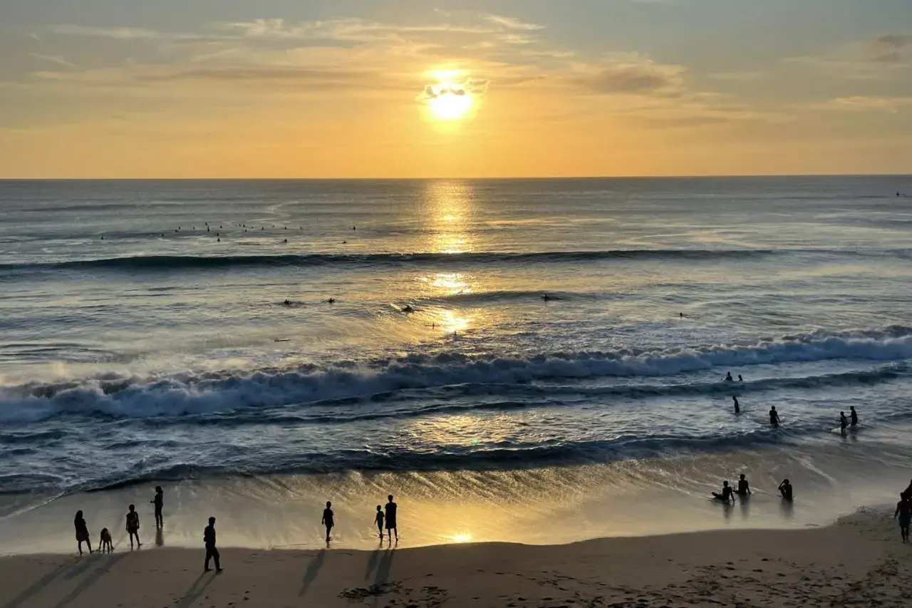 Zachód słońca na plażach Bali. Surferzy czekają na fale, a ludzie spacerują po piasku, ciesząc się złotym blaskiem.