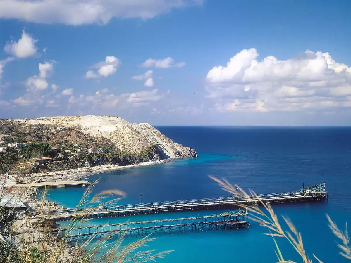 Panoramica delle isole Lipari con un porto e moli che si estendono nel mare blu intenso sotto un cielo azzurro con nuvole bianche.