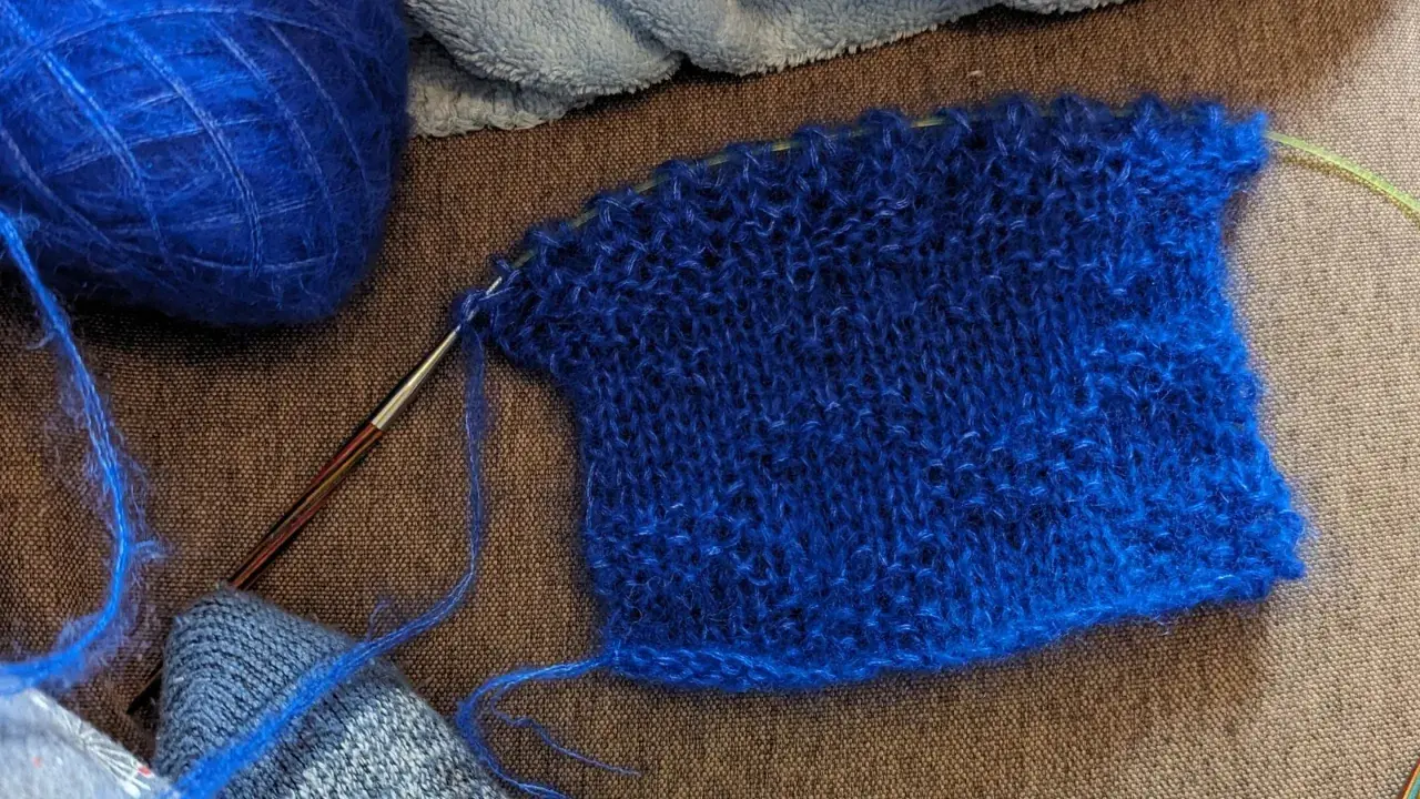 Un échantillon de tricot bleu vif, avec une pelote de laine assortie et une aiguille. La jauge maille est bien visible sur le tissu.