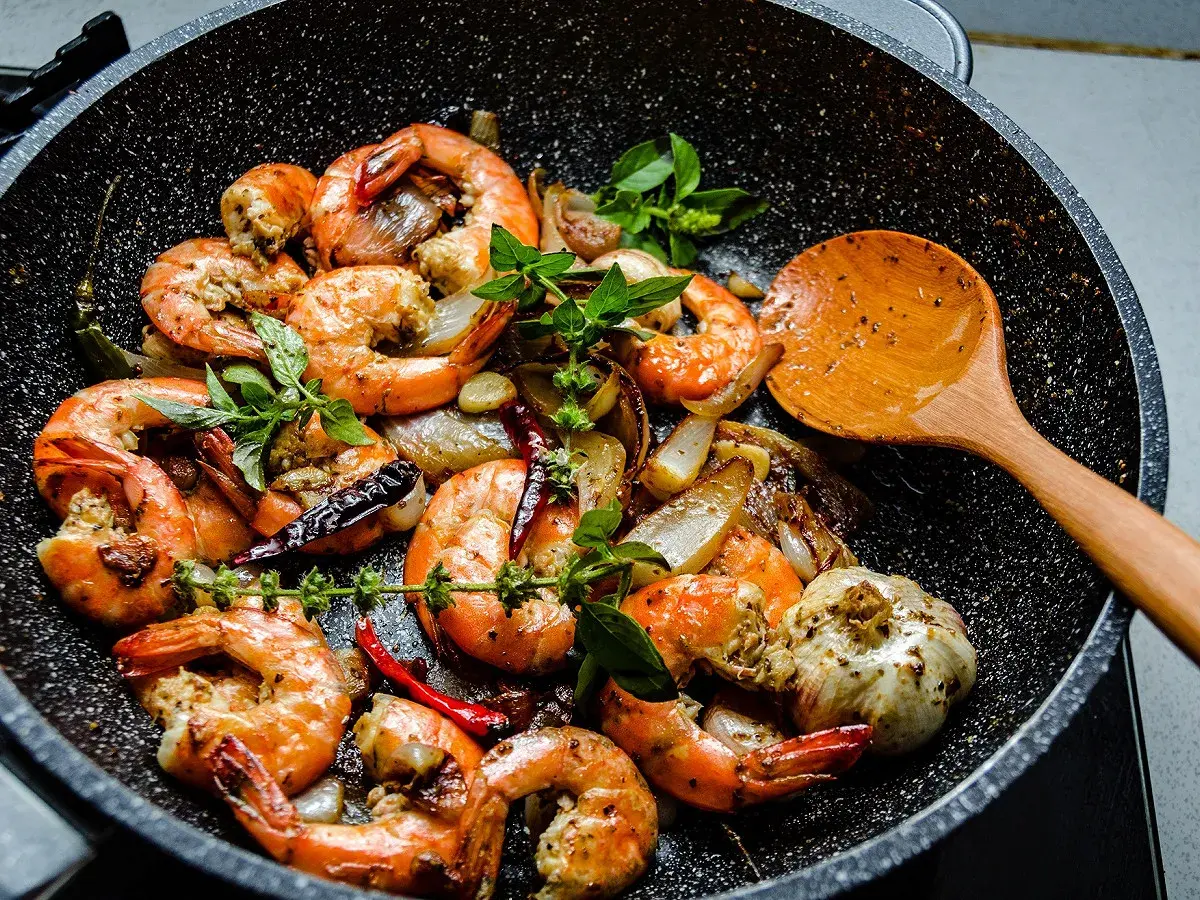 Gambas sautées à l'ail et au piment dans une poêle, prêtes pour le temps de cuisson. Une cuillère en bois est à côté.