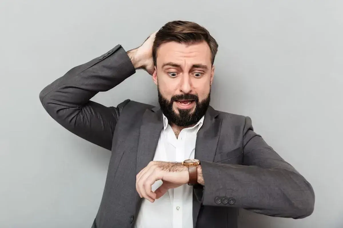 Hombre con traje gris, barba y expresión de pánico, se lleva una mano a la cabeza mientras mira su reloj. ¡Parece que va a soñar que llega tarde al trabajo!