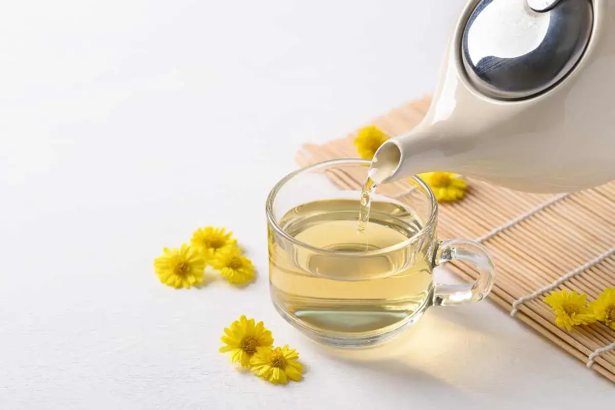 Tè giallo dorato versato da una teiera in una tazza trasparente, circondato da piccoli crisantemi fiori gialli.