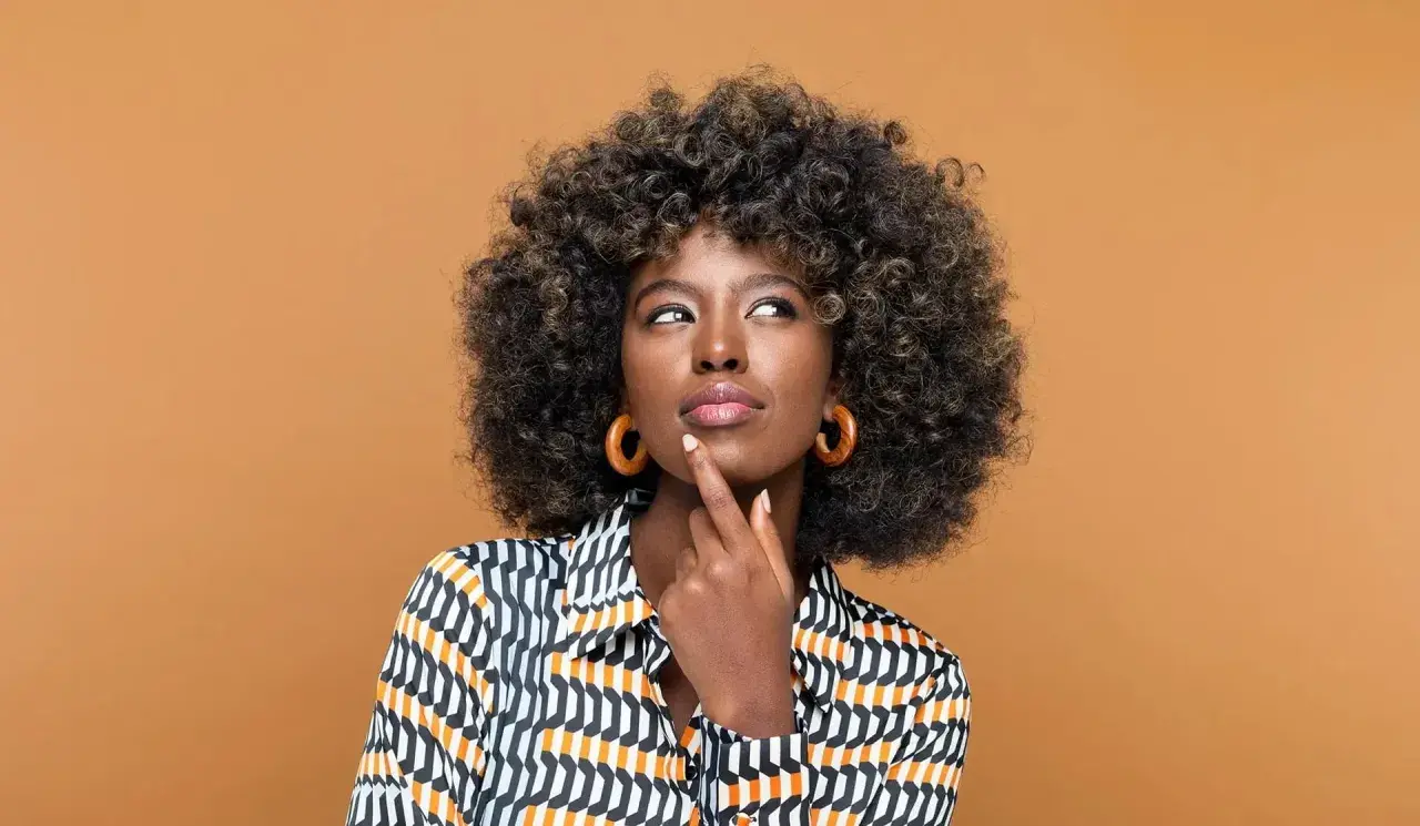 Mujer pensativa con cabello rizado y coloración semipermanente, luciendo una blusa estampada y aretes circulares.