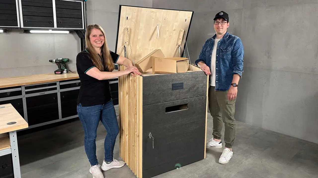 Ein Paar präsentiert stolz seine selbstgebaute Paketbox. Die Box ist aus Holz gefertigt und hat eine Klappe, die mit Ketten gesichert ist. Sie ist groß genug, um mehrere Pakete aufzunehmen.