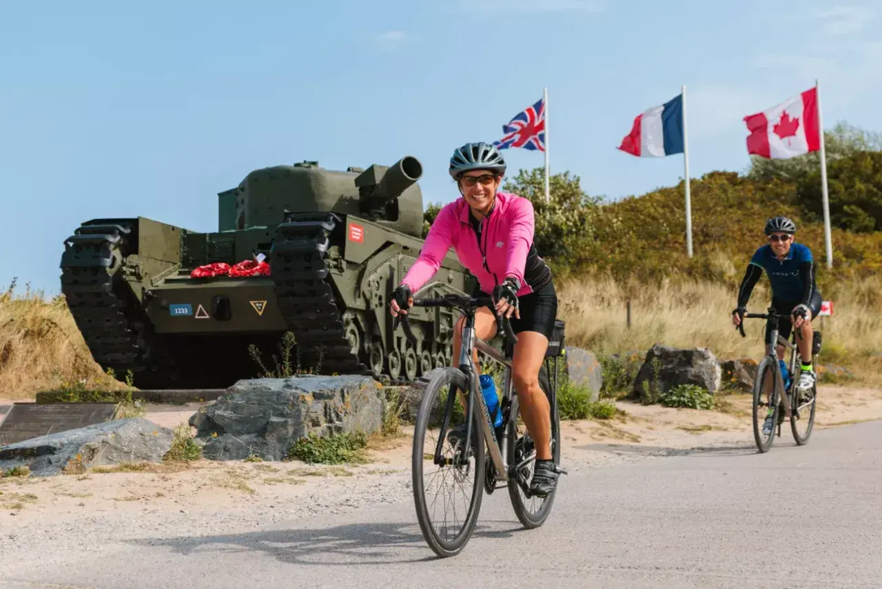Cyclists enjoy Normandy bike tours past a tank memorial with Union Jack, French, and Canadian flags flying.