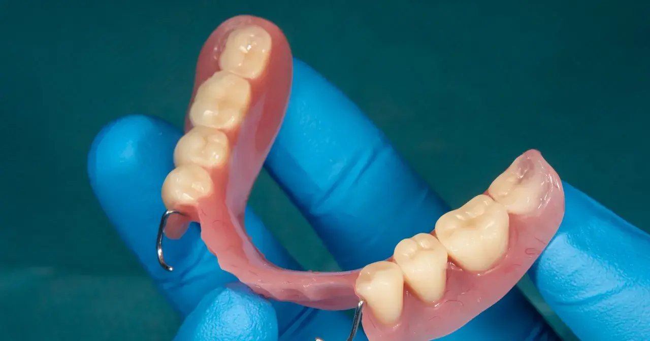 Proteza natychmiastowa na NFZ, wykonana z różowego akrylu z zębami, trzymana w niebieskich rękawiczkach.