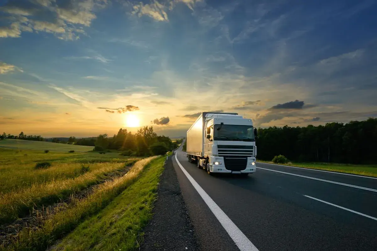 Un camion blanc roule sur une route au coucher du soleil, traversant une campagne verdoyante. C'est le quotidien de ceux qui travaillent sur la route.
