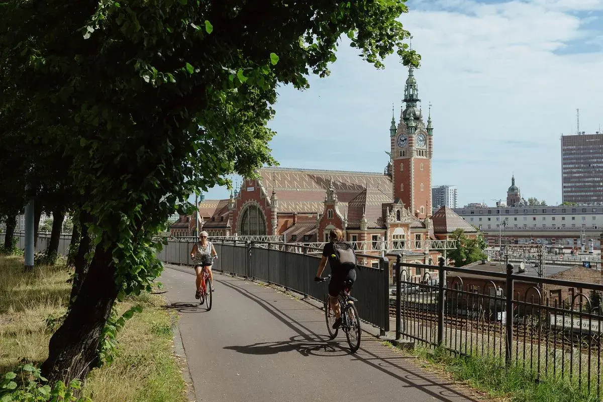 Dwie osoby na rowerach jadą ścieżką obok torów kolejowych. W tle piękny budynek dworca z wieżą zegarową. To idealne miejsce przyjazne rowerzystom.