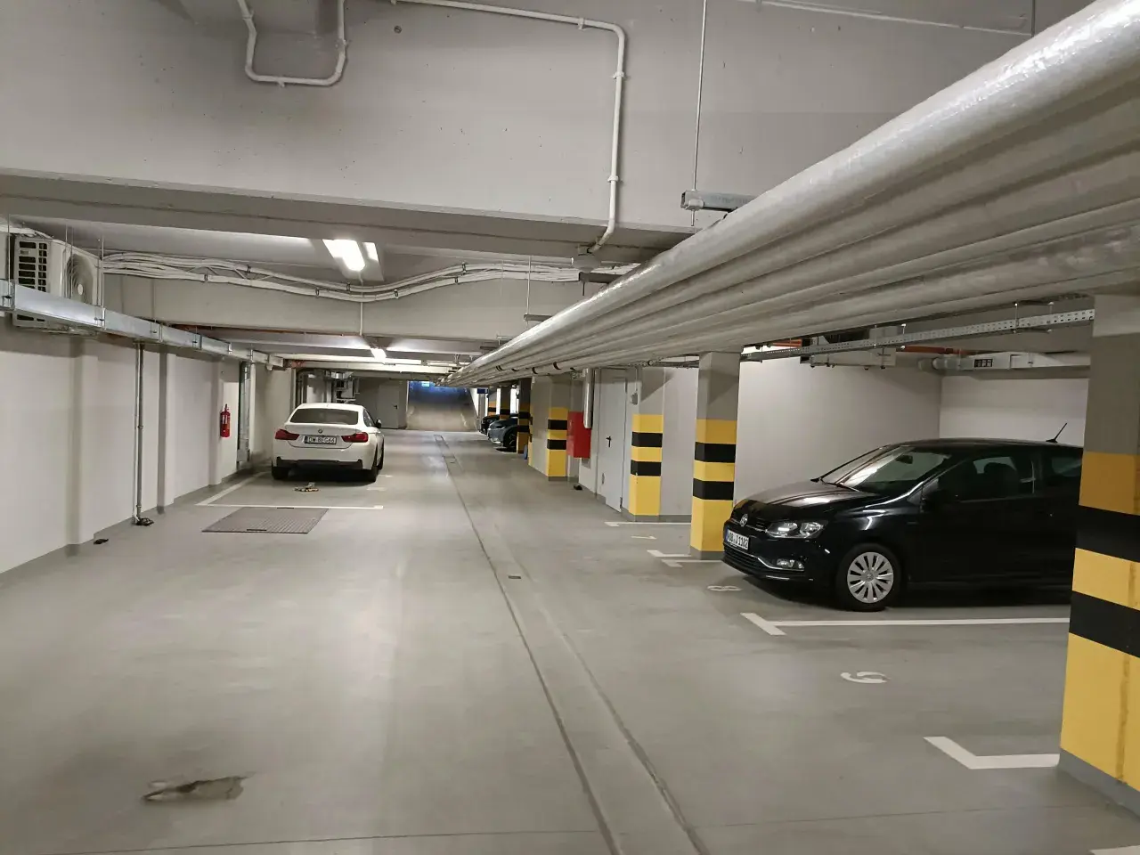 Wrocław główny parking cennik: podziemny parking z białym i czarnym Volkswagenem, białym BMW, rurami i klimatyzacją.