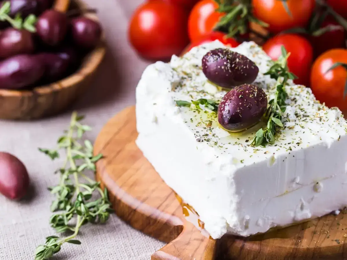 Un blocco di feta greca condita con erbe aromatiche e olive su un tagliere di legno, con pomodori e olive in sottofondo.