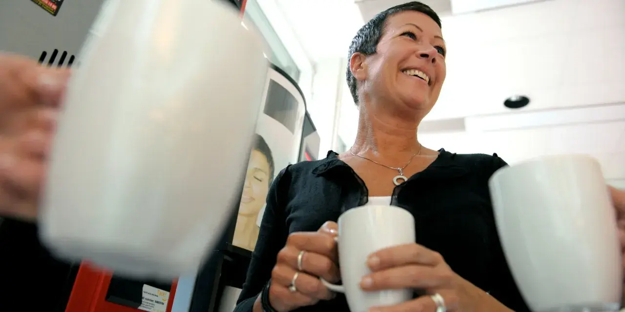 En kvinna ler glatt med en kaffemugg i handen. Hon verkar njuta av stunden, vilket visar vad betyder inflytande i vardagen.