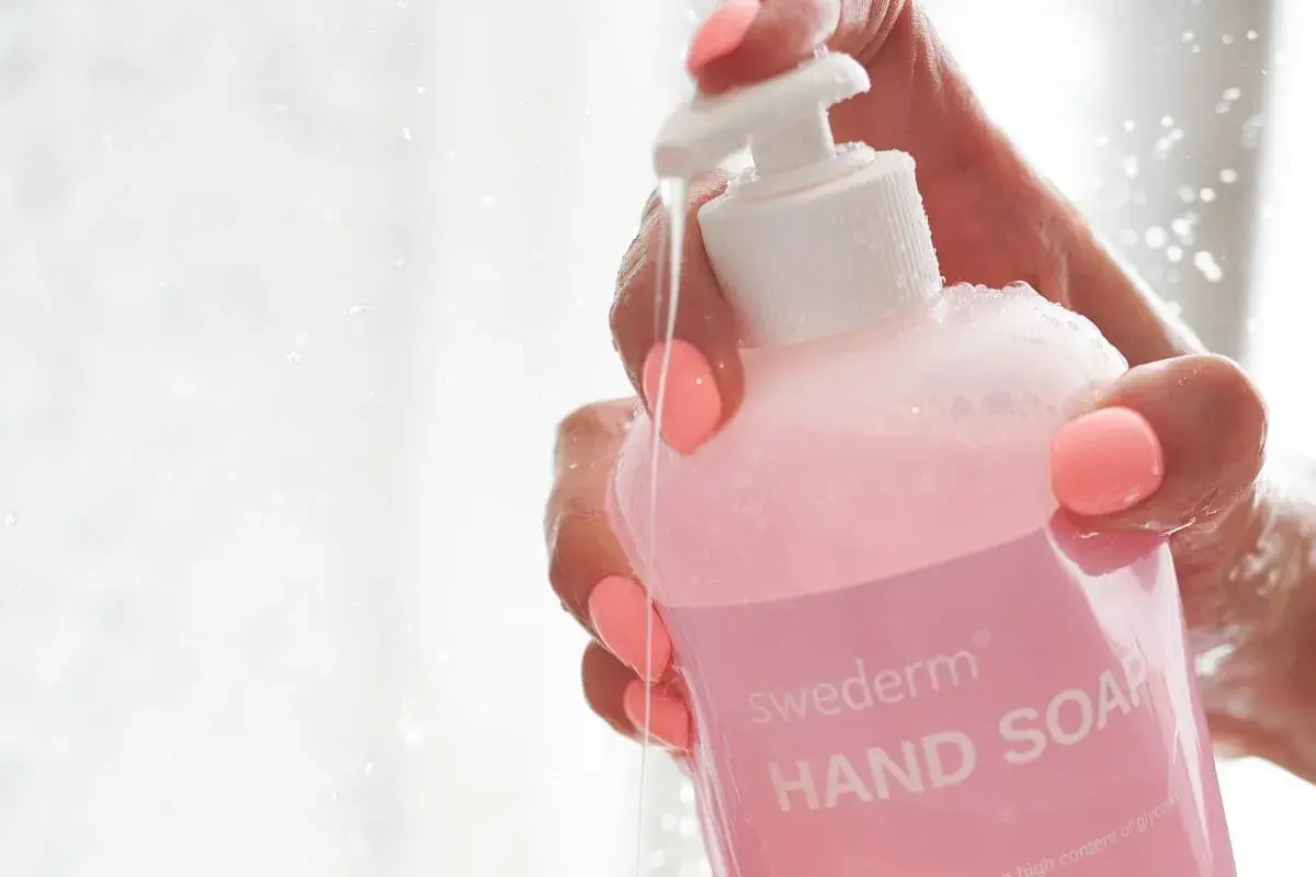Dłoń z różowymi paznokciami trzyma różowy płyn do mycia rąk Swederm. Woda spływa po butelce, a na etykiecie widać napis "HAND SOAP". Czy gliceryna jest szkodliwa? Ten produkt zawiera jej wysoki poziom.