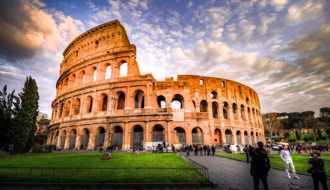Il Colosseo, un'icona di cosa visitare a Roma, si erge maestoso sotto un cielo drammatico. Turisti passeggiano sul selciato, ammirando l'antica grandezza.
