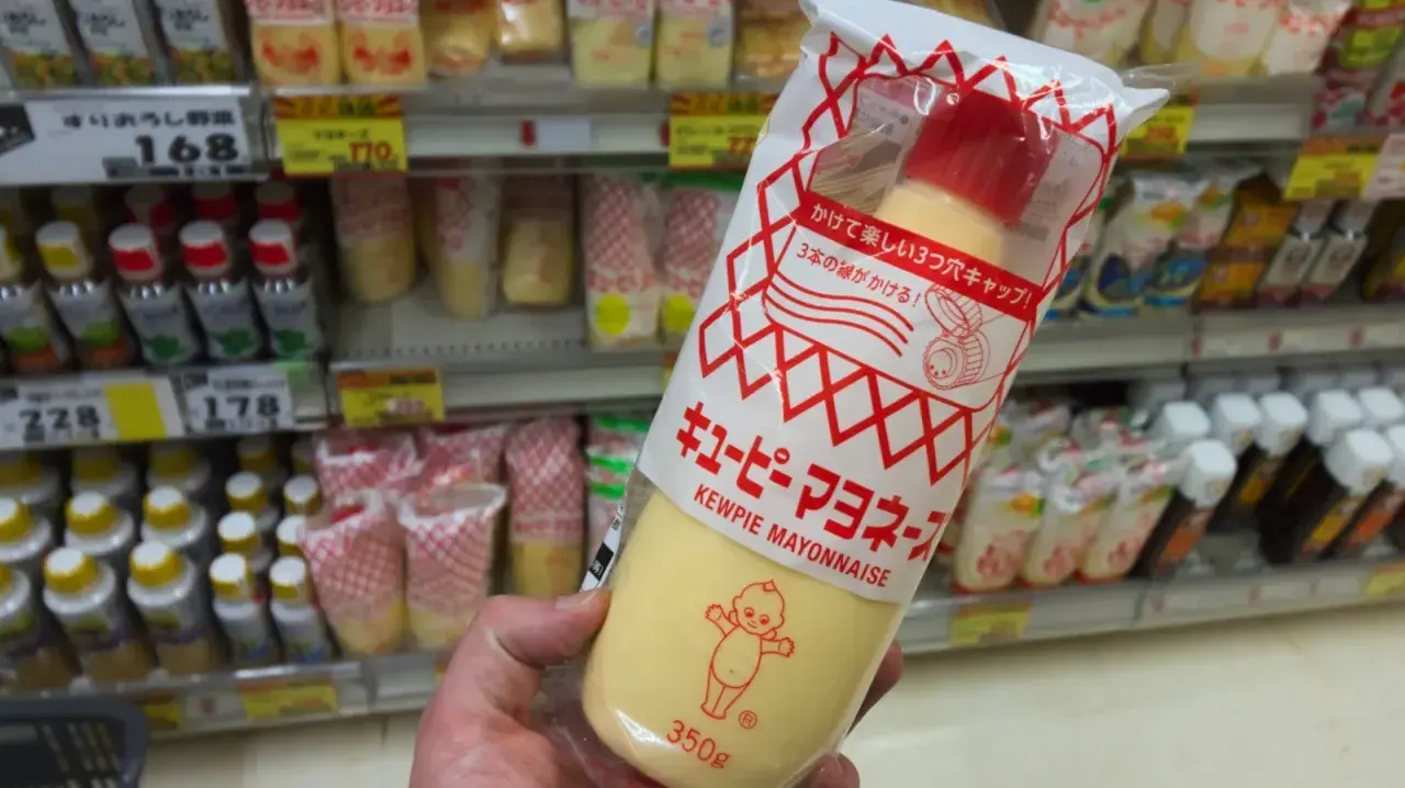 A hand holds a large squeeze bottle of Kewpie mayonnaise. The packaging features the iconic Kewpie doll. The question "how long does Kewpie mayo last?" is on my mind as I shop.