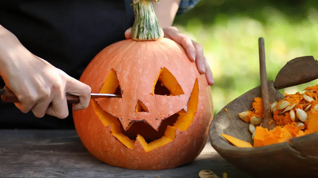 Händer skär ett ansikte i en pumpa för en klassisk pumpasminkning. Bredvid ligger en skål med pumpafrön och fruktkött.
