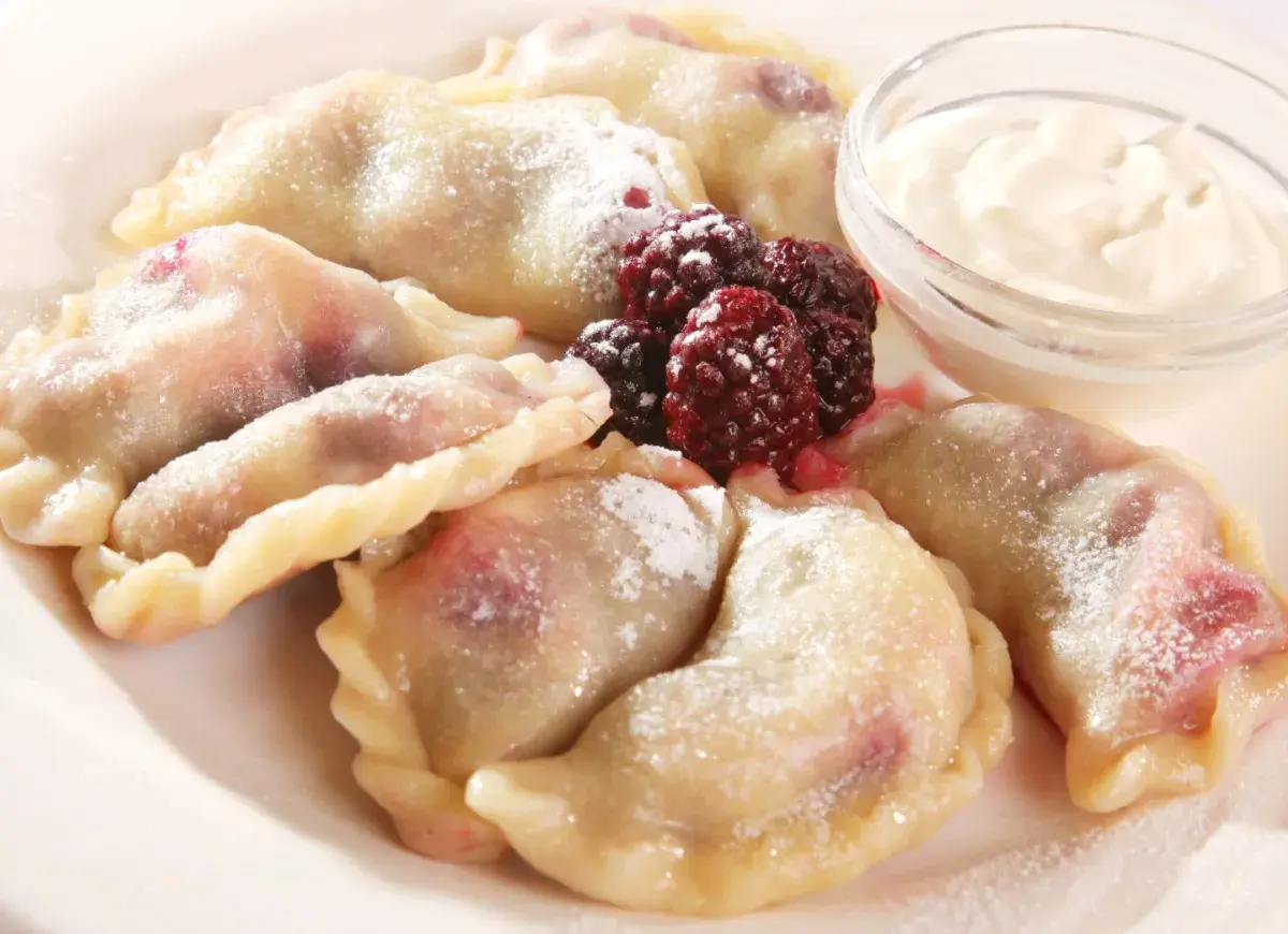 Pyszne pierogi z malinami, posypane cukrem pudrem, podane z kwaśną śmietaną i świeżymi owocami leśnymi.