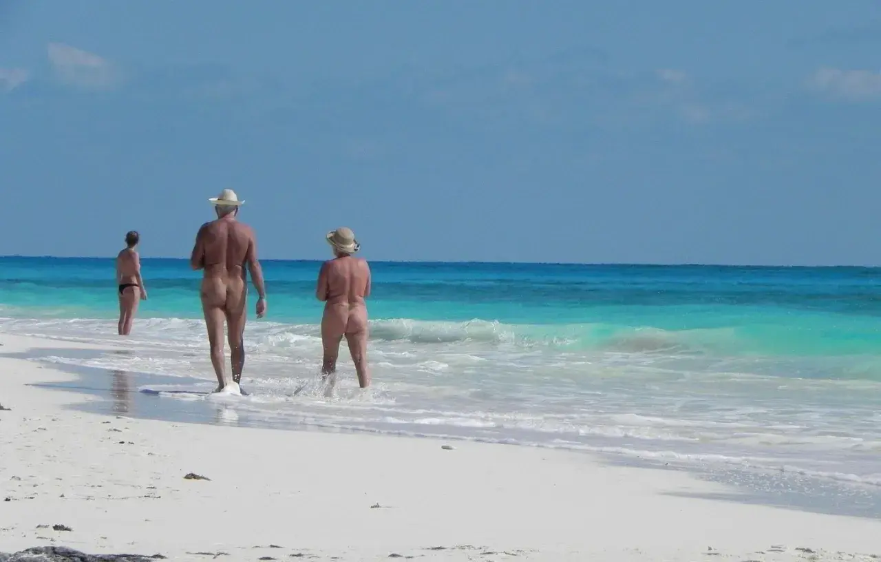 Trois personnes profitent d'une plage italienne, l'une en bikini, les deux autres nues, dans un décor de camping naturiste.