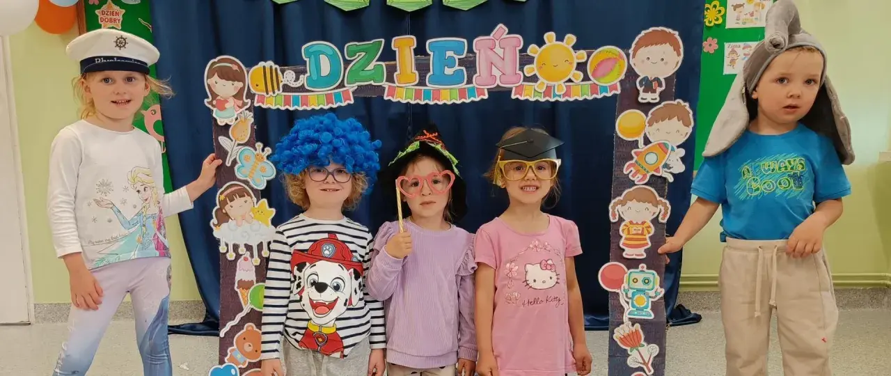 Dzieci w przedszkolu w strojach i z rekwizytami pozują do zdjęcia na tle dekoracji z napisem "Dzień Dziecka".