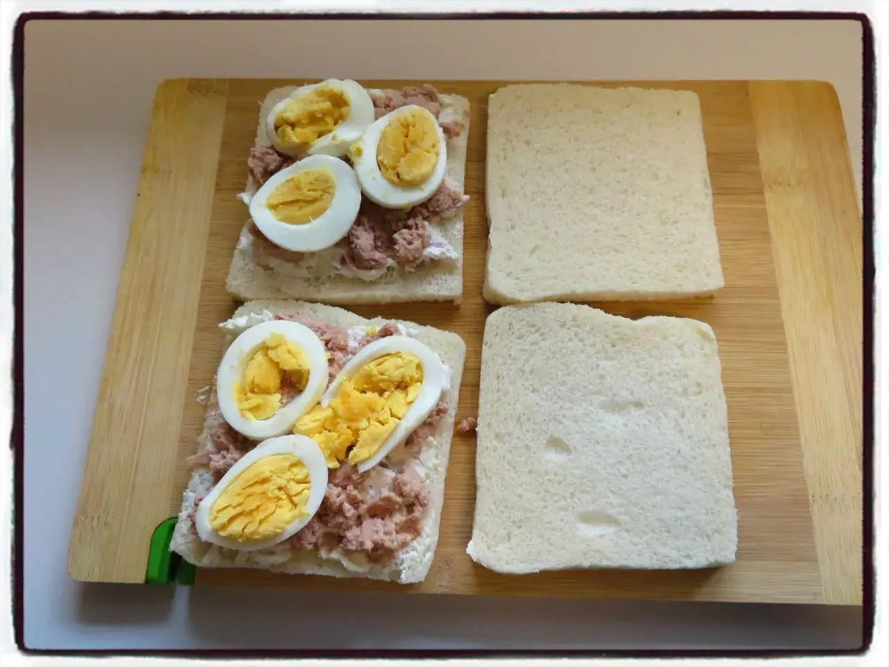 Deux sandwichs oeuf dur sont en préparation sur une planche en bois. Le thon émietté et la mayonnaise sont déjà étalés sur le pain.