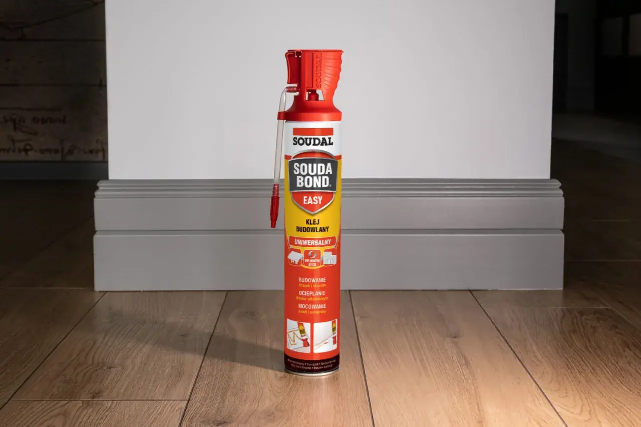 Puszka kleju budowlanego Soudal Easy Bond na drewnianej podłodze, obok szarej listwy przypodłogowej.