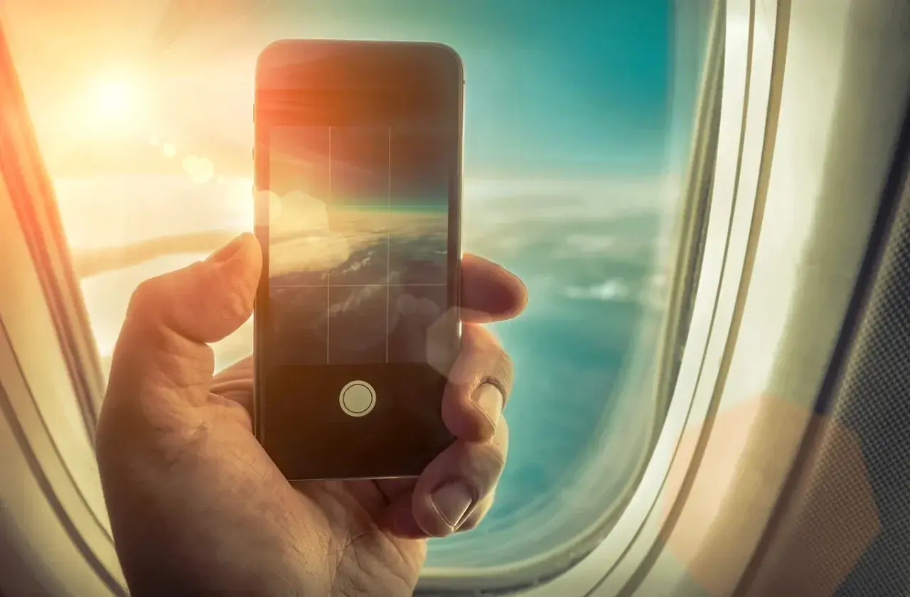 Dłoń trzyma smartfon, którym fotografuje chmury z okna samolotu. Czy w samolocie można używać telefonu? Tak, do robienia zdjęć!