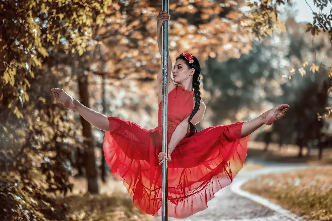 Kobieta w czerwonej sukience wykonuje figurę na słupie, jak podczas kursu pole dance. Jej nogi są rozłożone w szpagacie, a włosy splecione w warkocz.