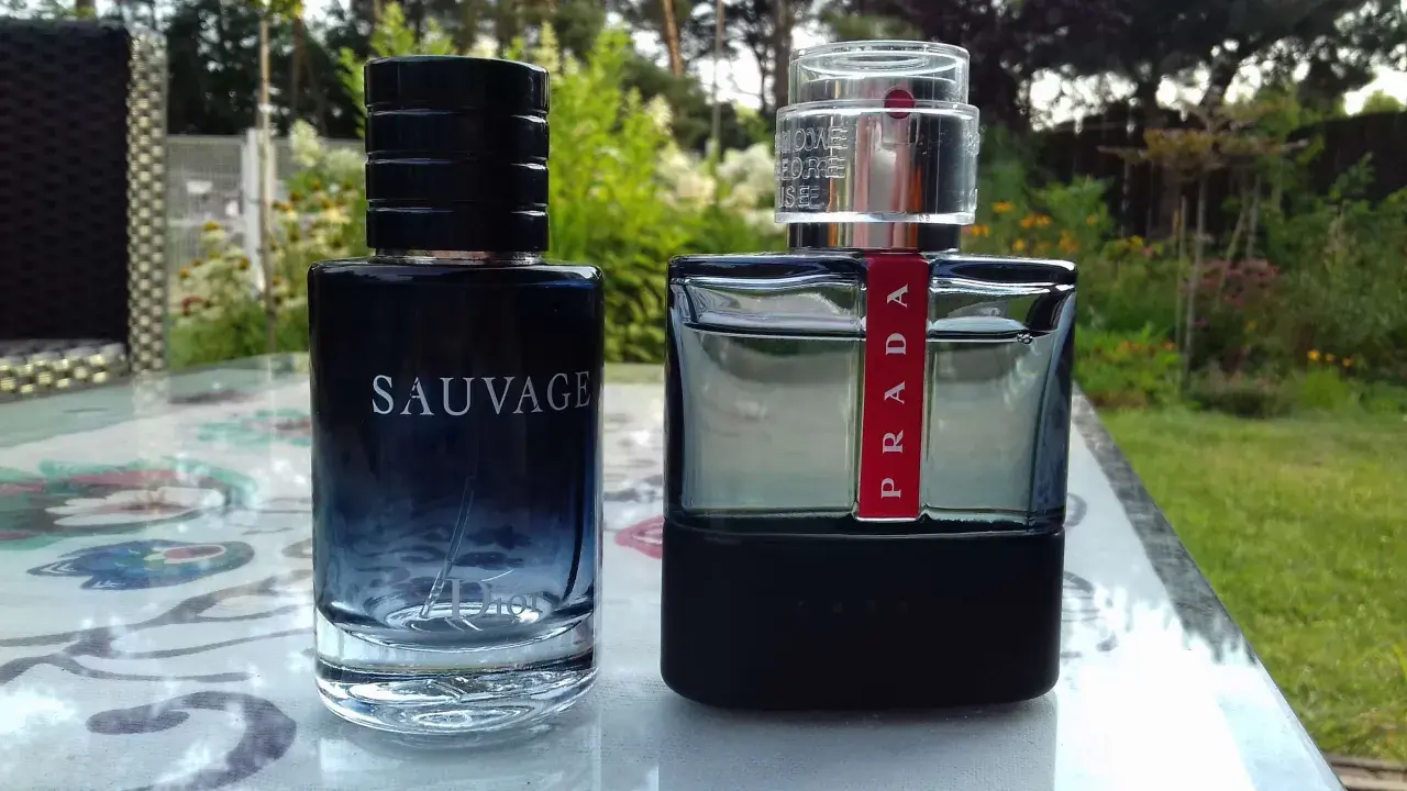 Dwie butelki perfum: Dior Sauvage i Prada Carbon, stoją na stoliku w ogrodzie.