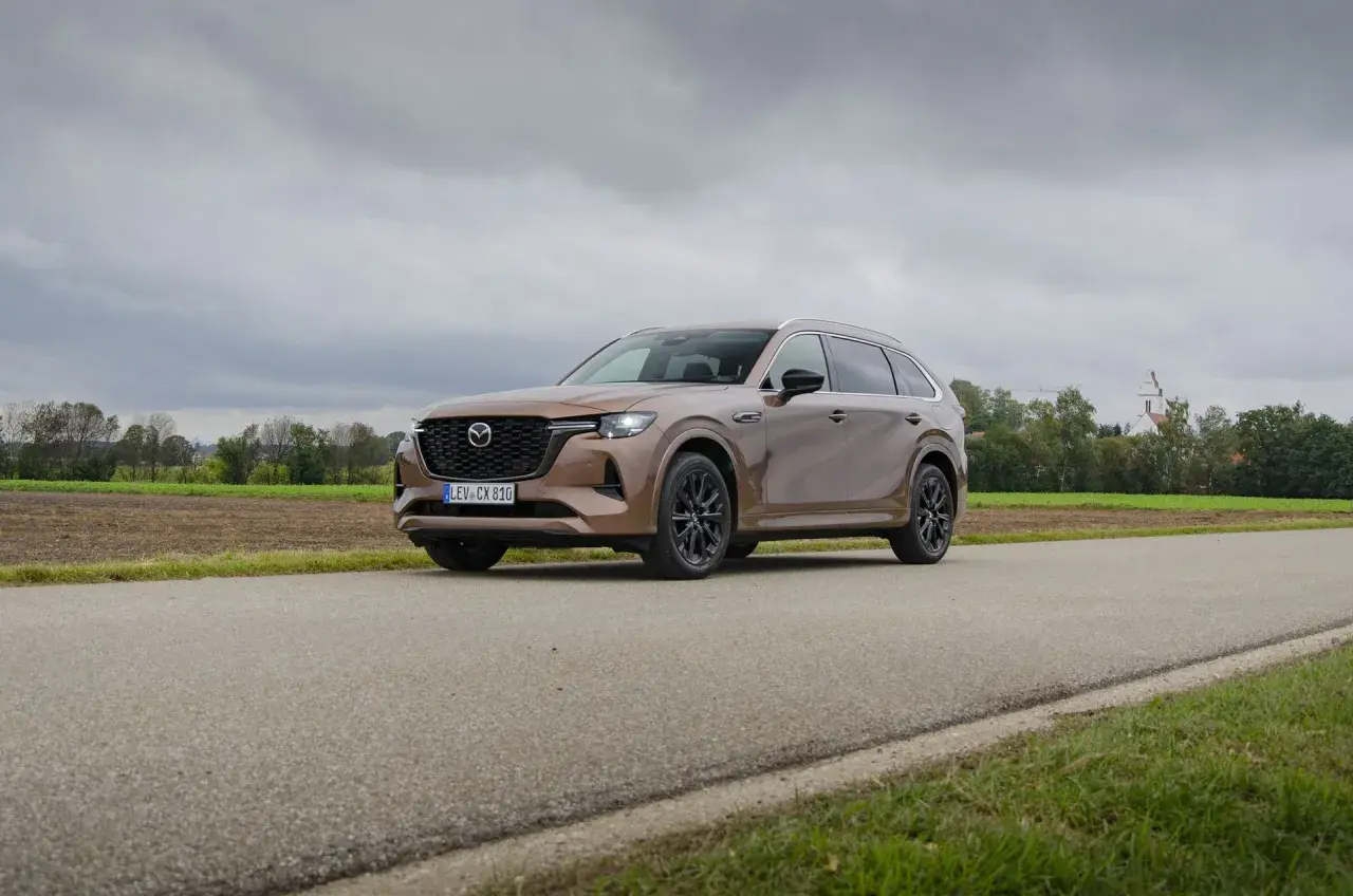 Brązowa Mazda CX80 na tle wiejskiego krajobrazu z kościołem w oddali.