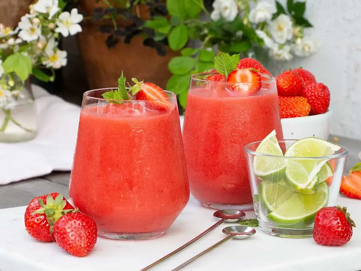 Två frostiga jordgubbsdaiquiri-drinkar med mynta och jordgubbar, serverade med limeklyftor och färska jordgubbar.