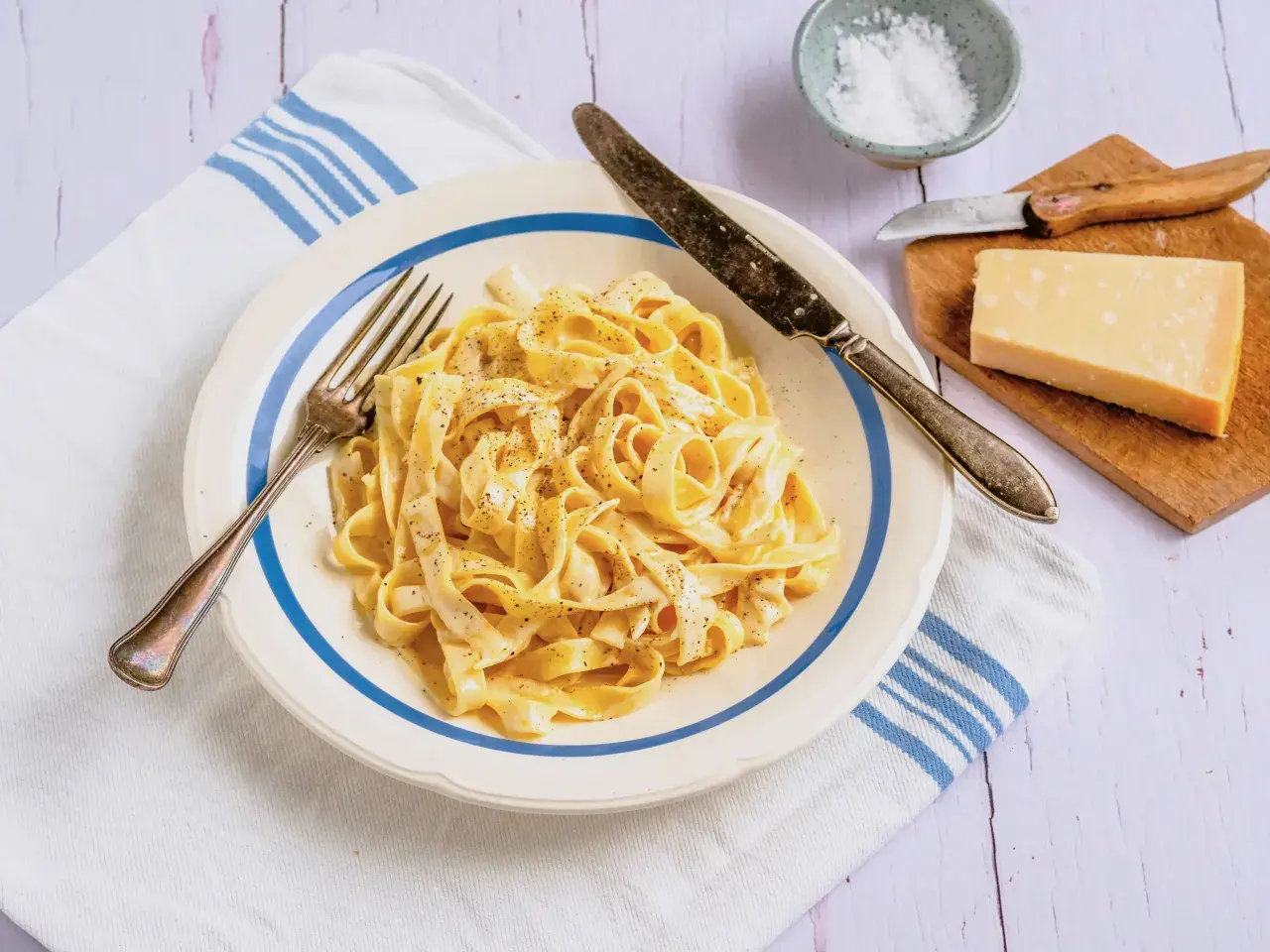 Ein Teller cremiger Fettuccine, bestreut mit schwarzem Pfeffer, daneben ein Stück Parmesan und Salz.