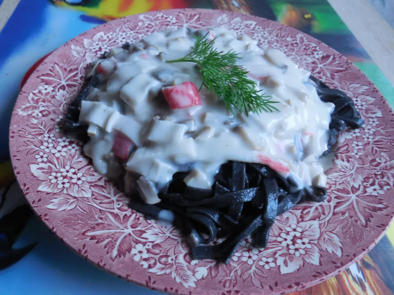 Czarny makaron z kremowym sosem i kawałkami kraba, udekorowany gałązką koperku. Pyszne danie na różowym talerzu.