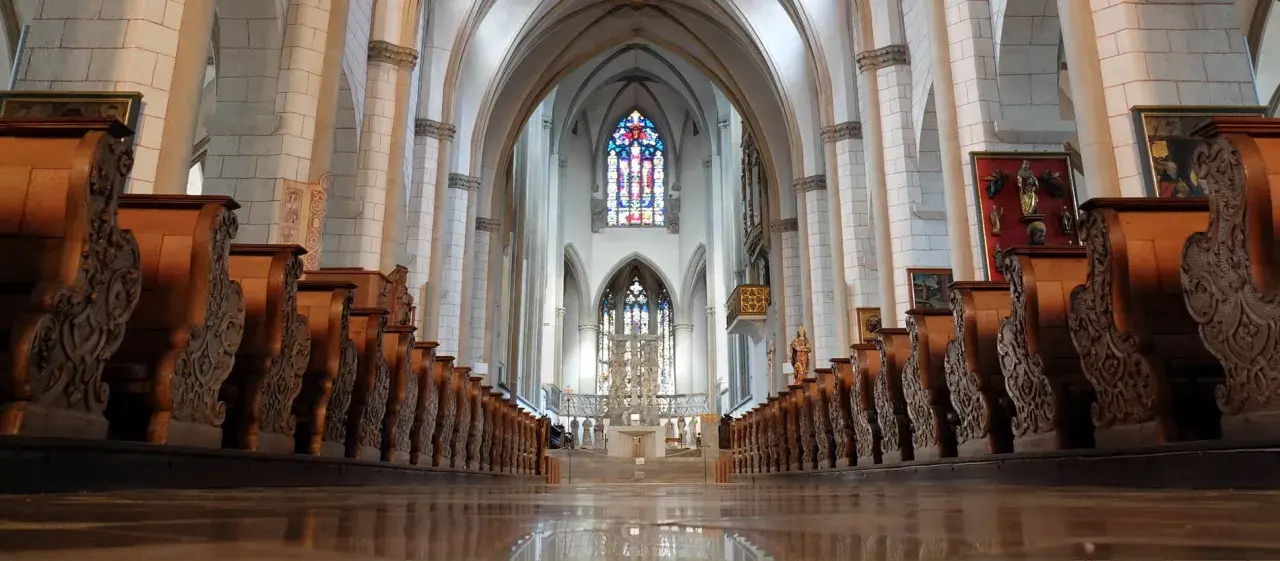 Blick durch das Kirchenschiff des Augsburger Doms mit seinen kunstvoll geschnitzten Holzbänken und dem farbenprächtigen Chorfenster.